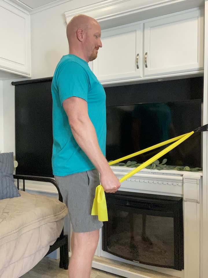 Coach Todd, a fitness professional, demonstrating pulldowns with a resistance band for a person with rounded shoulders by pulling the band towards their chest while standing with their feet shoulder-width apart and the band anchored above them.