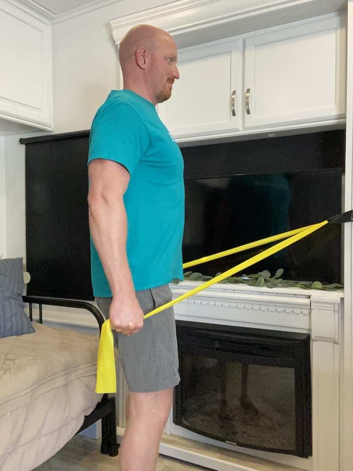 Coach Todd, a fitness professional, demonstrating pulldowns with a resistance band for a person with rounded shoulders by pulling the band towards their chest while standing with their feet shoulder-width apart and the band anchored above them.
