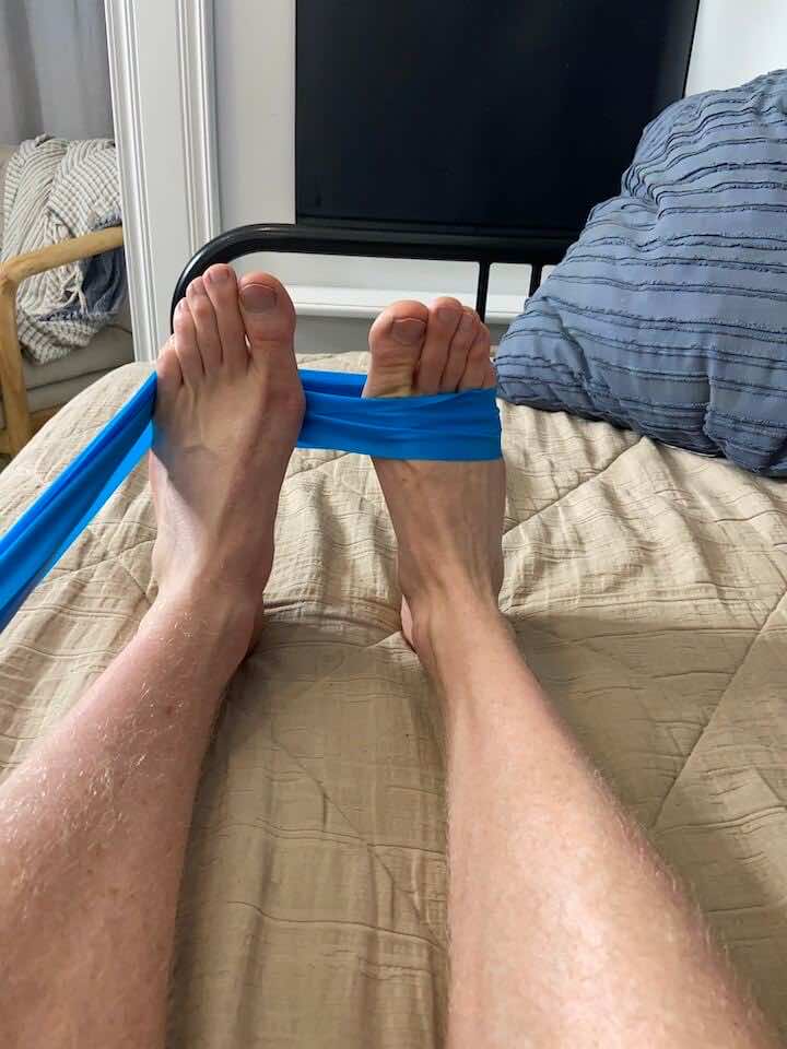 Lay down on your bed with your ankle off the end. Loop the band around your right foot and around your left. We will use your left foot as an anchor for your right foot as your exercise. Bring your right foot out like you're angling your ankle away from your left foot, squeeze in, then relax. ankle eversion with resistance band 1